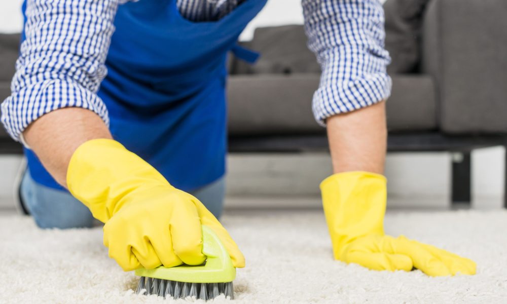 Nettoyage manuel d'un tapis avec une brosse par une personne portant des gants jaunes.